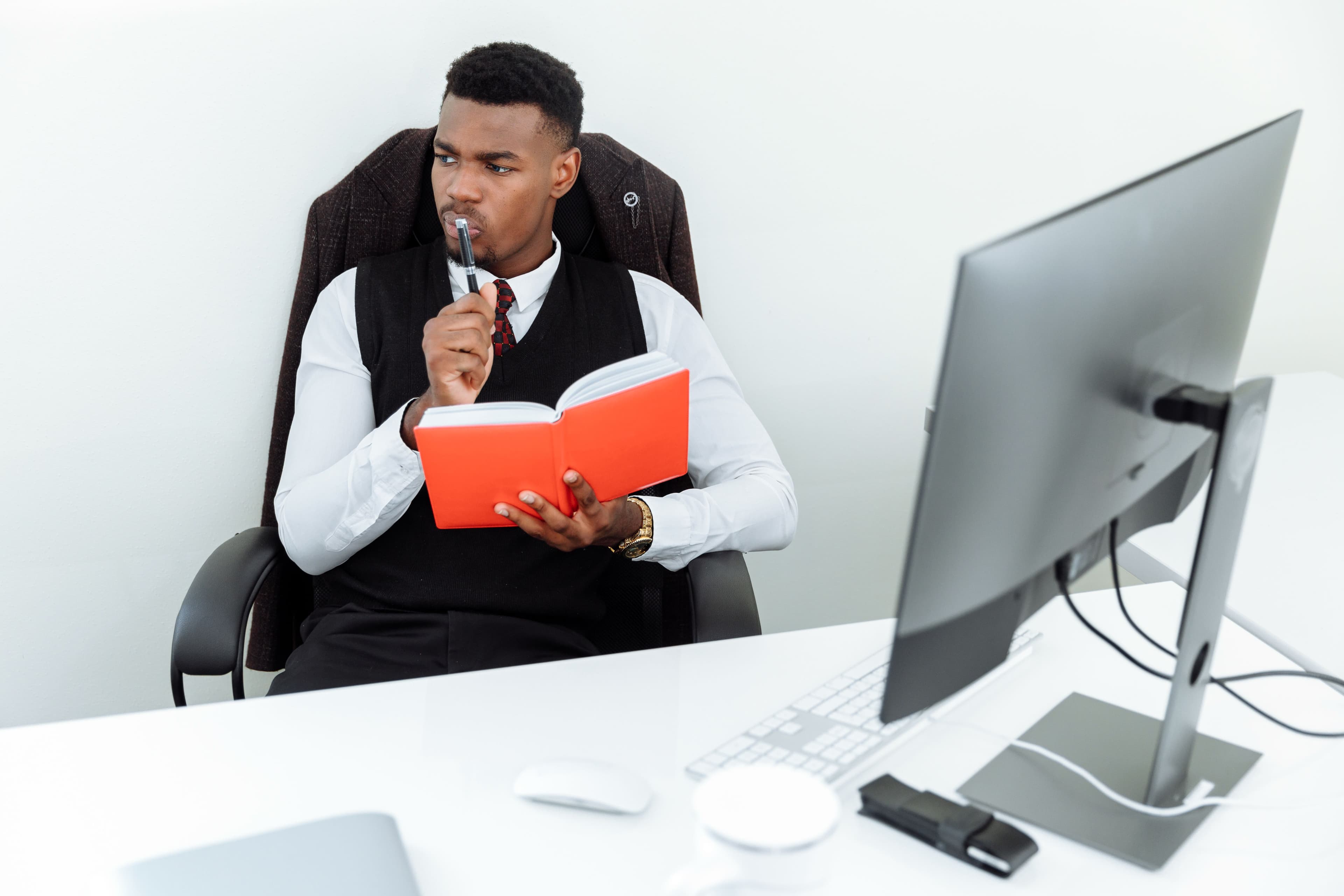 A man in a desk chair writing in a notebook beside a large monitor.